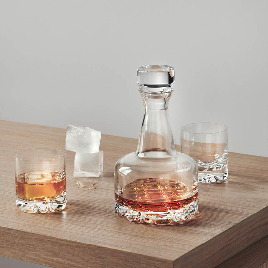 A clear decanter and two Orrefors: Erik Double Old Fashioned Glasses 12oz from a 4-pack, filled with amber liquid, sit on a wooden surface next to three ice cubes.