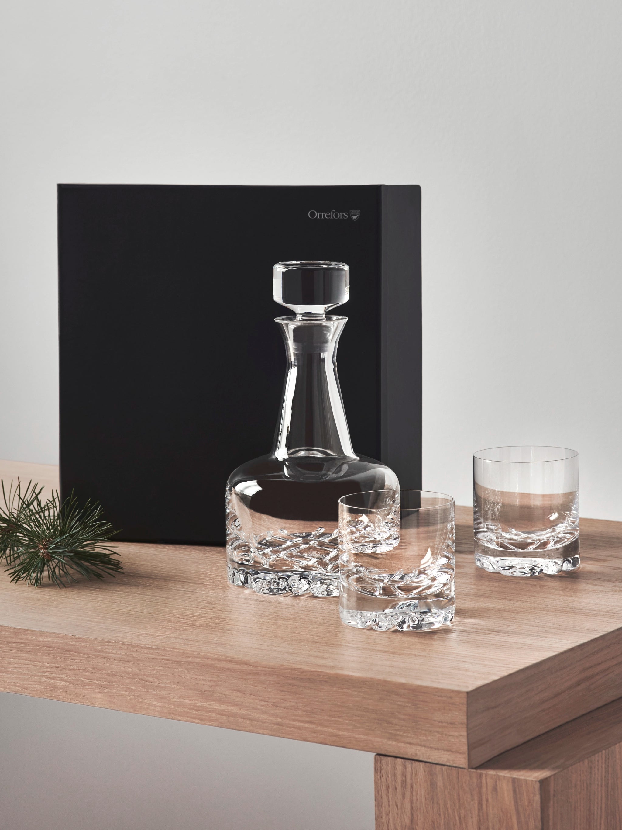 The Orrefors Erik 3 Piece Set, featuring a clear glass decanter and two Double Old Fashioned glasses, sits elegantly on a wooden table. A small pine branch and a black box complete the serene background.