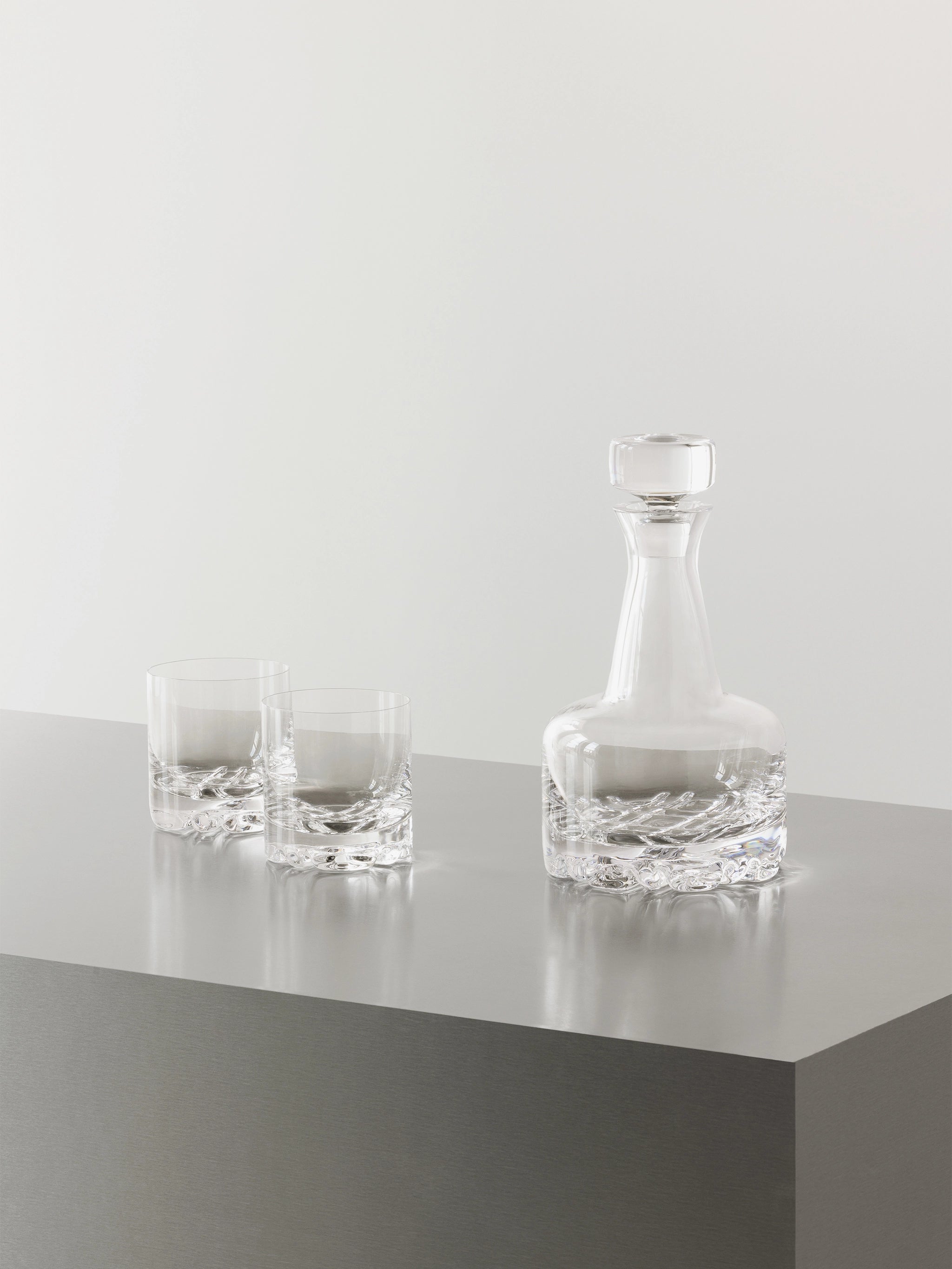 The Orrefors: Erik 3 Piece Set includes a clear glass decanter and two matching DOF glasses, beautifully displayed on a smooth, gray surface against a plain white background.