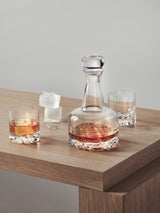 The Orrefors Erik 3-Piece Set includes a 26oz glass decanter filled with whiskey, placed on wood next to two 12oz double old fashioned glasses and three ice cubes, reflecting timeless Scandinavian design.