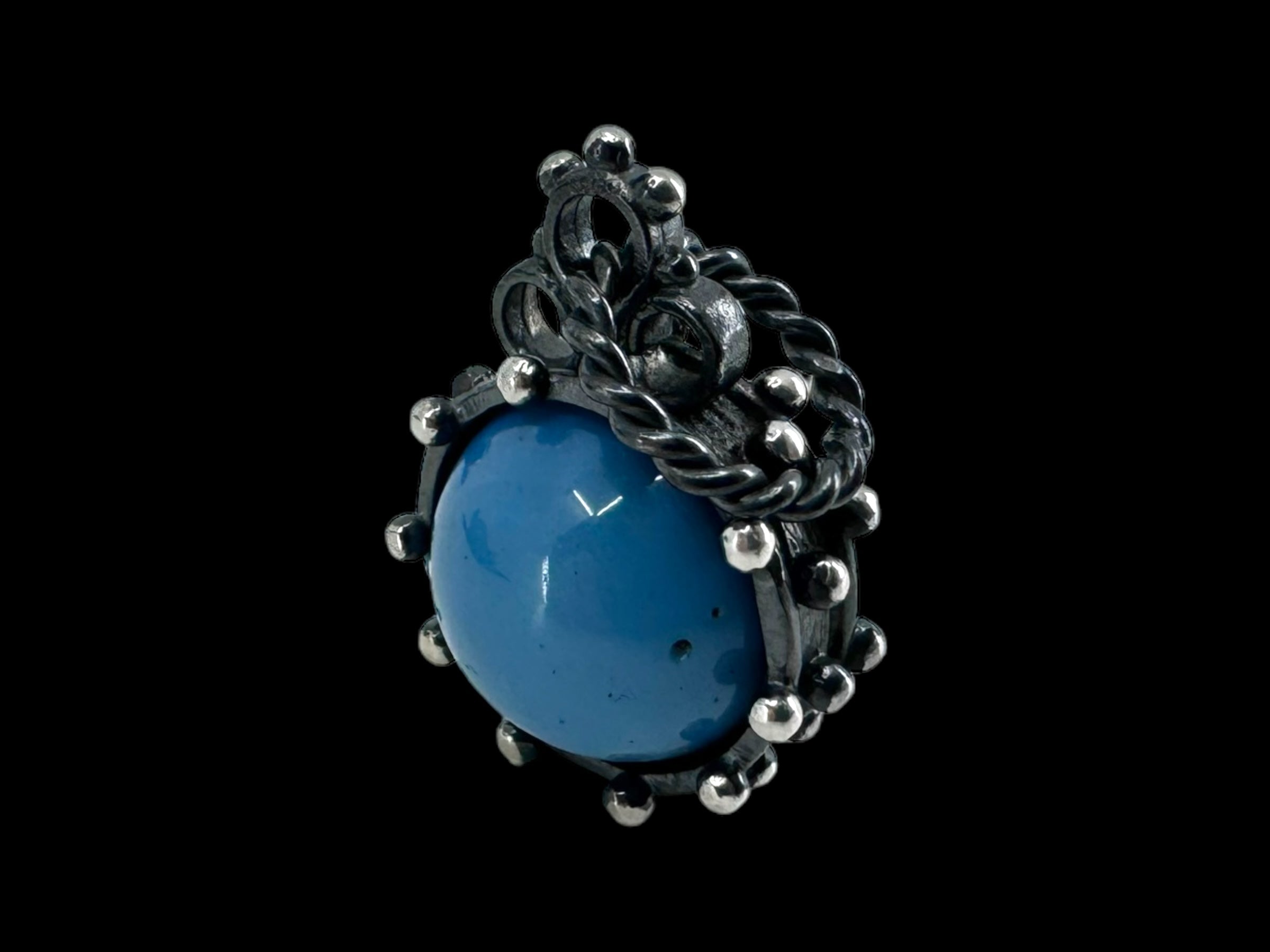 The Pendant: Freja - Swedish Blue features an eye-catching oxidized silver design with a large Swedish Blue stone at its center. It is embellished with delicate metalwork and tiny silver spheres, all set against a black backdrop.