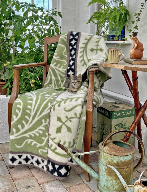 A kitten cozily nestles on an Albertus - 100% Norwegian Lambs Wool blanket by Kerstin Landstrom, draped over a wooden chair. Nearby, potted plants, a watering can, and a cup on a small table add to the charm and tranquility of the scene.