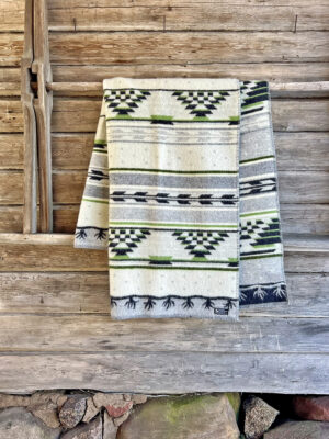 The Blanket: Sundborn Green, featuring geometric patterns in green, black, and gray and made from organic wool, is draped over a rustic wooden fence.