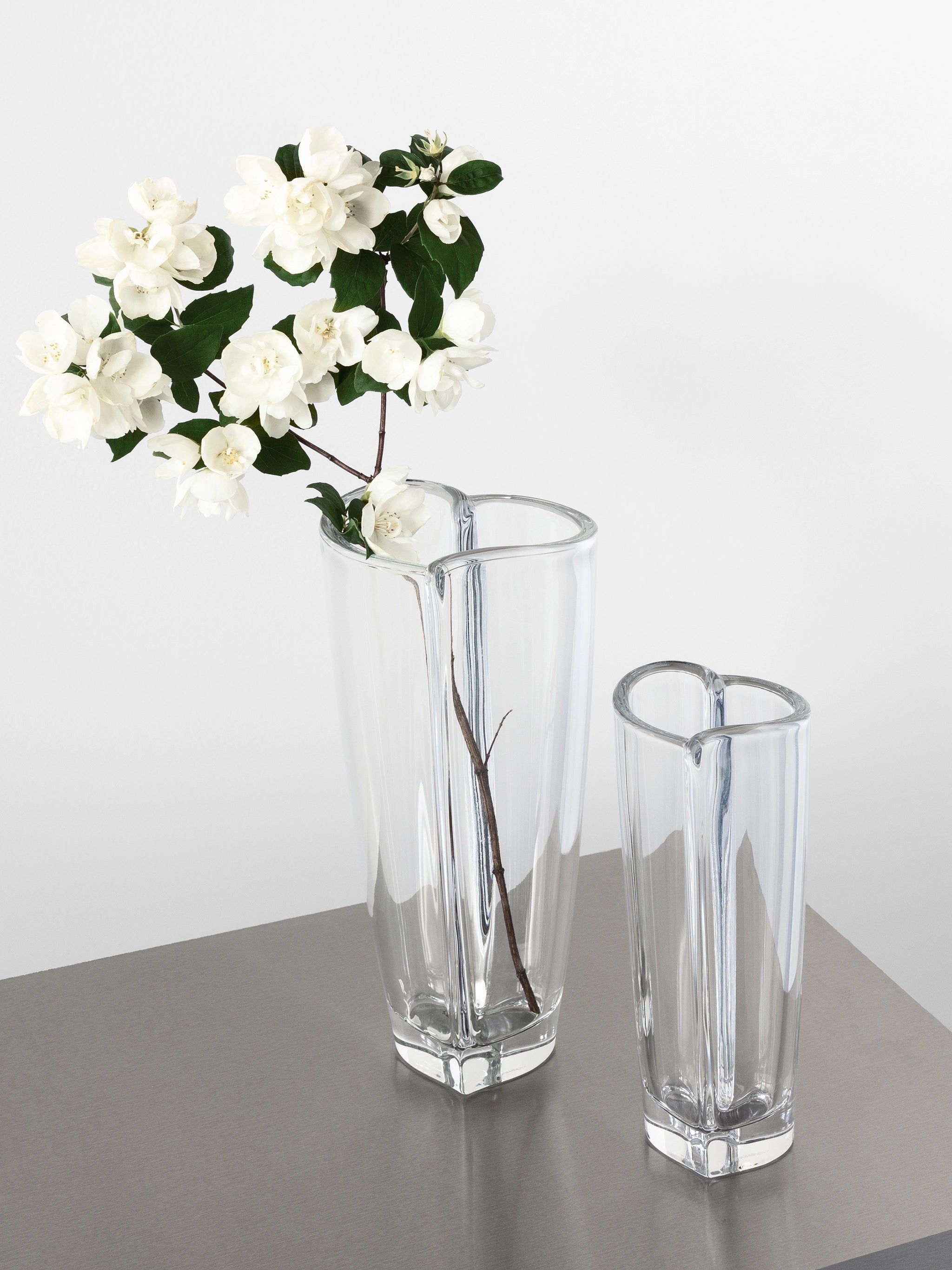 On a table sit two elegant vases; the taller Orrefors crystal glass vase cradles a branch with white flowers, while the Orrefors Heart Bud Vase remains empty.