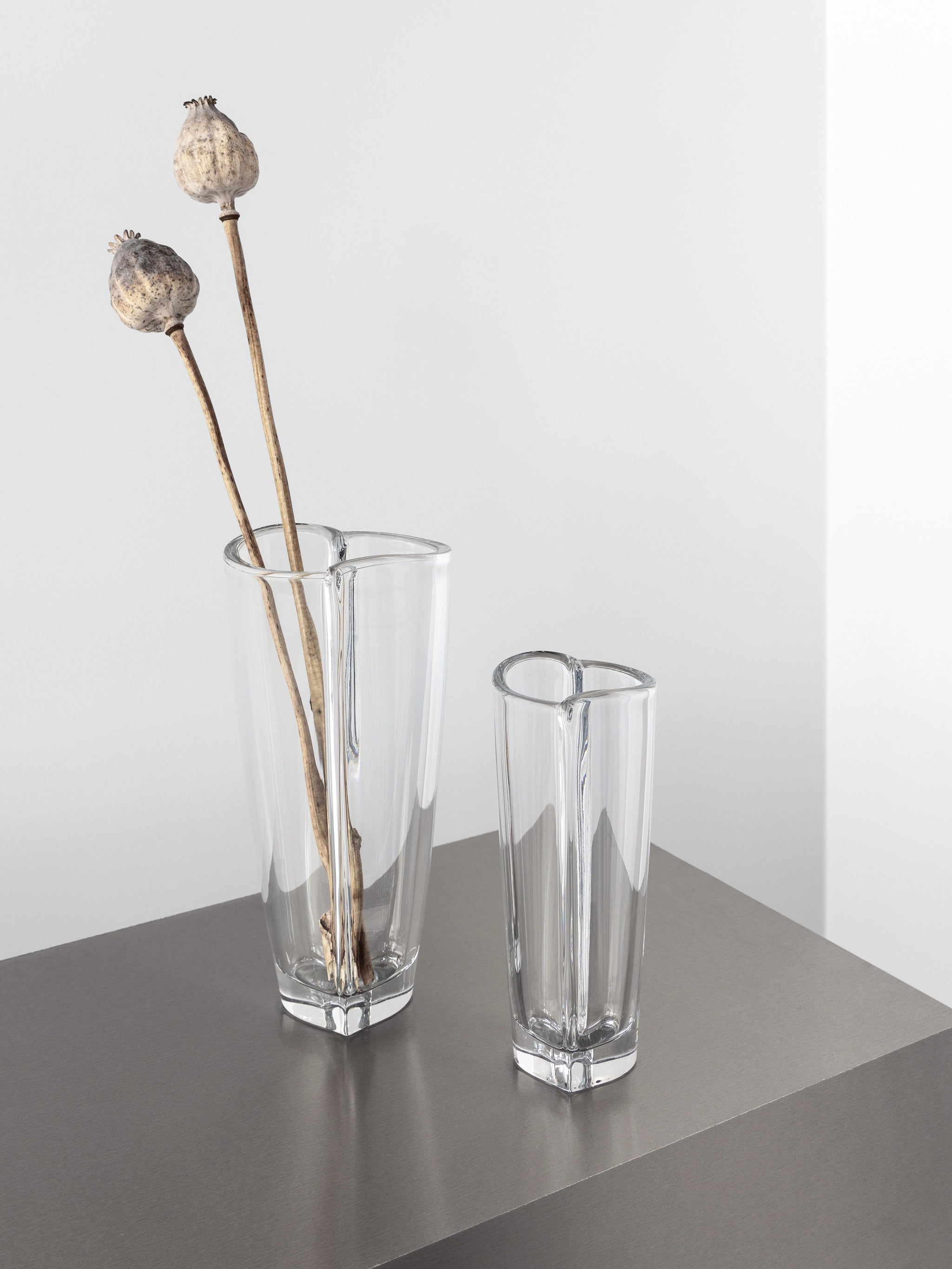 Two Orrefors Heart Bud Vases sit on a gray surface, with the larger one holding two dried poppy seed pods.