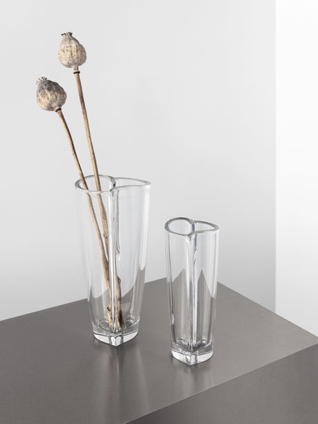 Two Orrefors Heart Bud Vases sit on a gray surface, with the larger one holding two dried poppy seed pods.