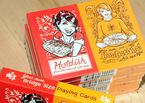 Several colorful boxes of Playing Cards: Hotdish/YouBetCha Bridge Size Playing Cards are stacked on a wooden surface, featuring illustrated women holding hotdish and pie.