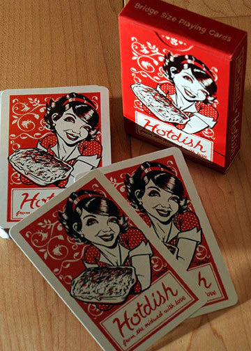 A red box of "Hotdish/YouBetCha Bridge Size Playing Cards" by Midwest sits on a wooden surface, with three cards visible—each featuring a smiling woman holding a dish.