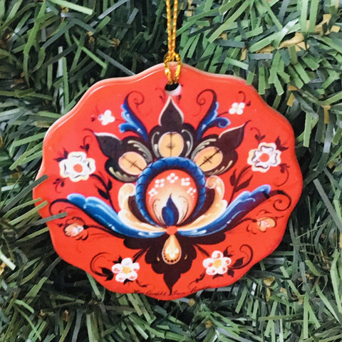 The Christmas tree is adorned with a Rosemaling Lise Lorentzen Ceramic Ornament, featuring a red floral porcelain design accented with shades of blue, brown, and white, adding unique charm to the festive display.