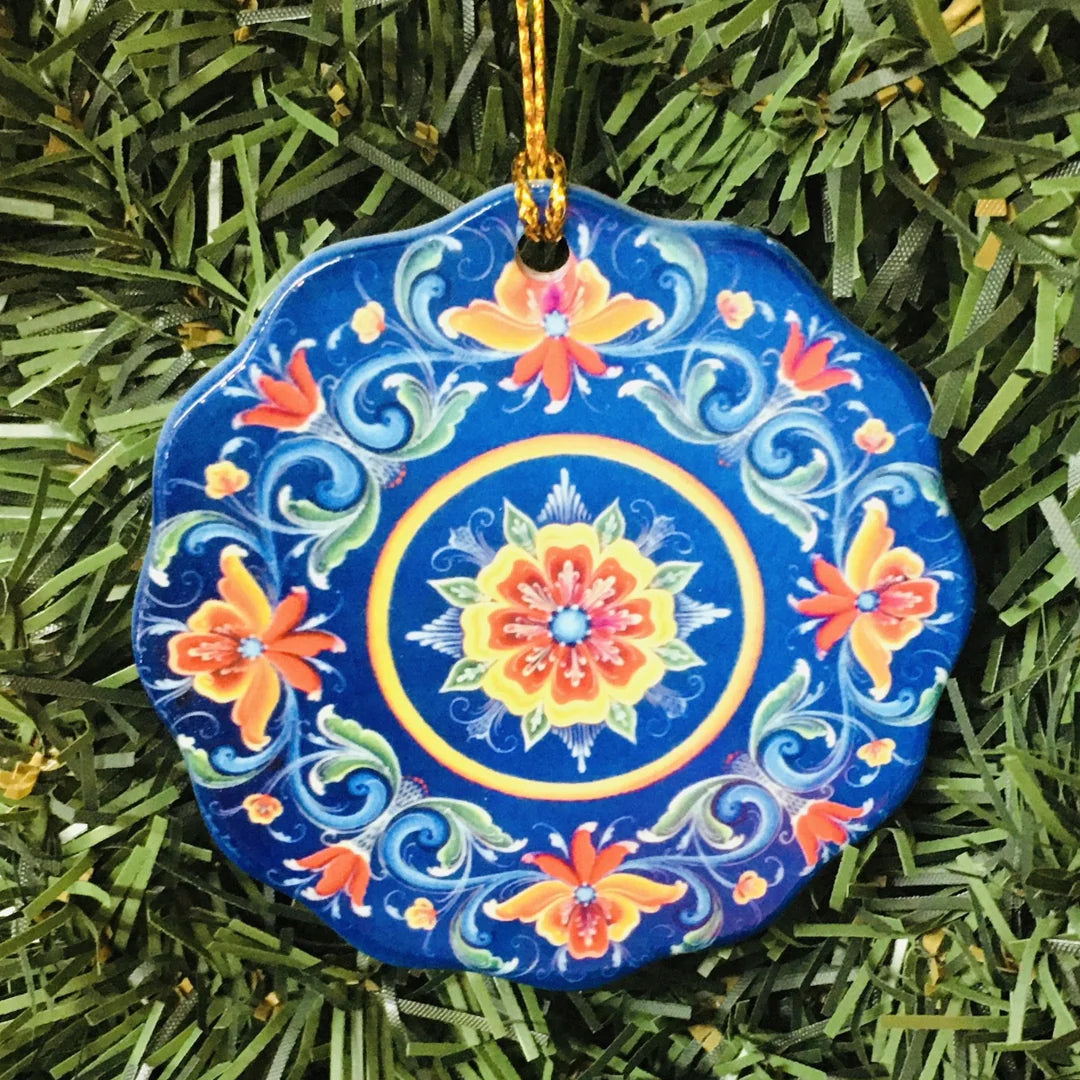 The Ornament: Lisa Lorentzen Ceramic Blue Rosemaling Ornament, featuring colorful floral patterns, hangs from a gold string on a green Christmas tree.
