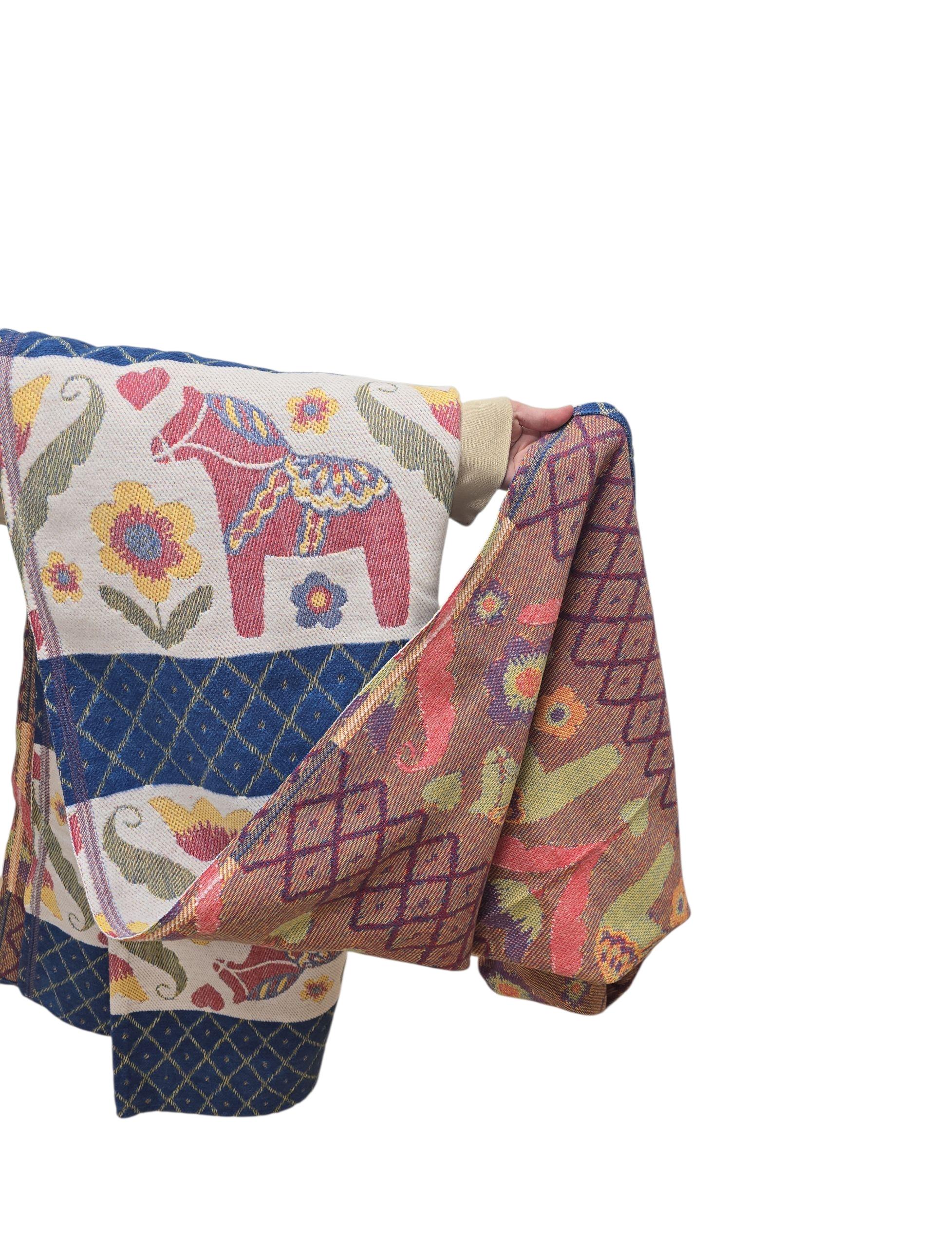 A hand holds up the Dala Horse 100% Cotton Woven Blanket by Ekelund of Sweden (55"x75"), featuring colorful floral and horse motifs, against a plain white background.