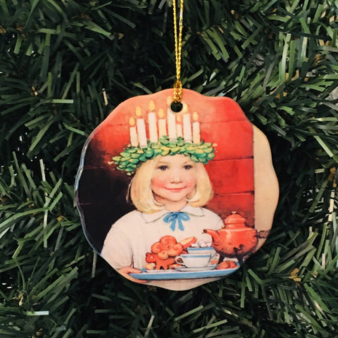 This porcelain ornament, named "St Lucia with Cinnamon Buns," captures the unique charm of a girl adorned with a candle crown. She delicately holds a tray featuring gingerbread cookies and a teapot, making it an ideal addition to your decoration project. It hangs beautifully on the green tree.