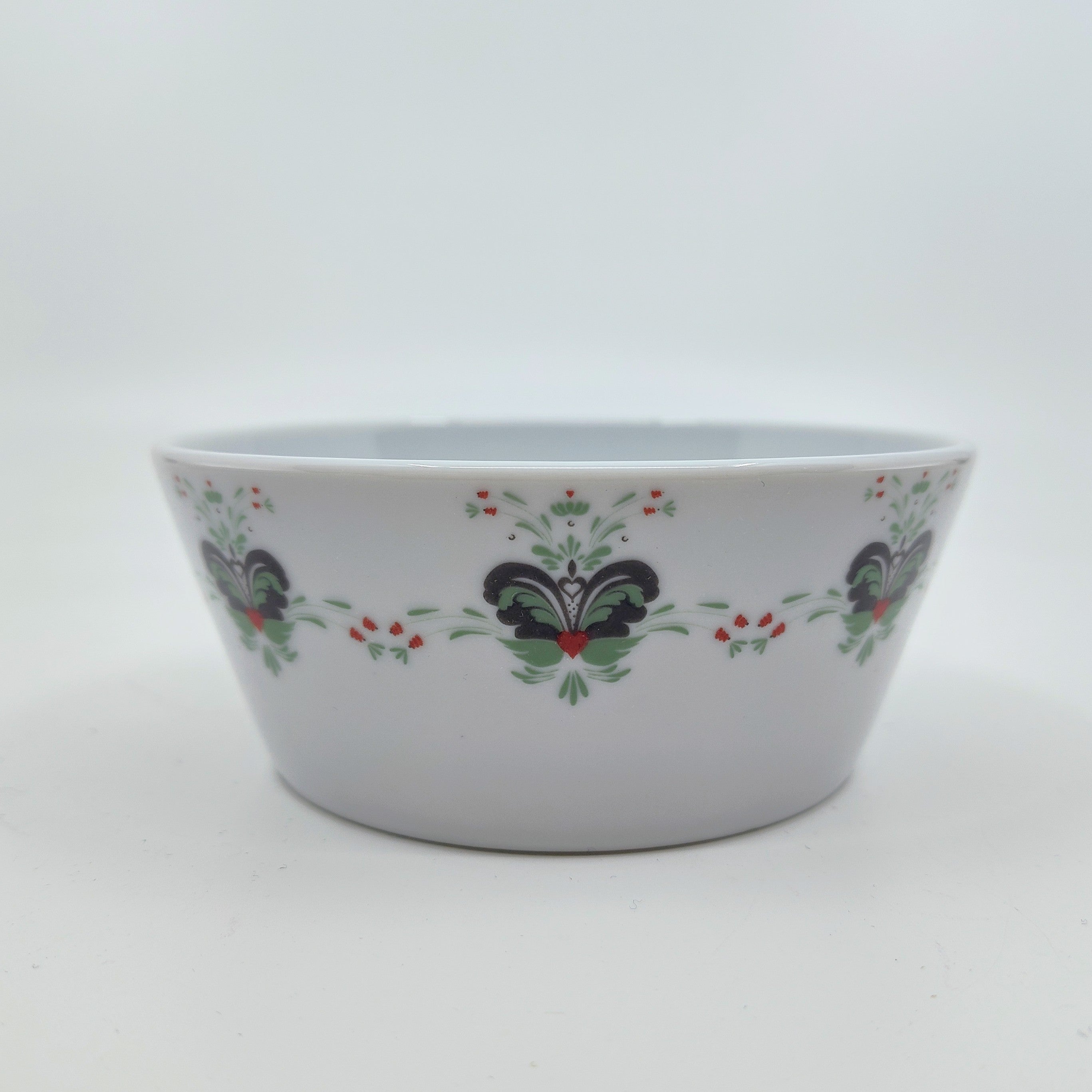 The Anna Viktoria Porcelain Bowl, adorned with a floral kurbits design in light green, grey, and red accents, is made in Portugal. It sits elegantly on a plain white background, highlighting its timeless charm.