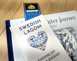 Three items on a wooden surface: the book “Swedish Lagom,” the bookmark “Swedish Proverbs ‘Life & People’” (2.5" × 8.5”), and “The Winter Journey” with an illustrated snowy scene on its cover.