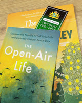 Three books are stacked on a wooden surface. The top book is titled The Open-Air Life, and a Bookmark: Set of 4 Swedish Proverbs (2.5" × 8.5") marks a page in the second book below, highlighting Scandinavian wisdom.