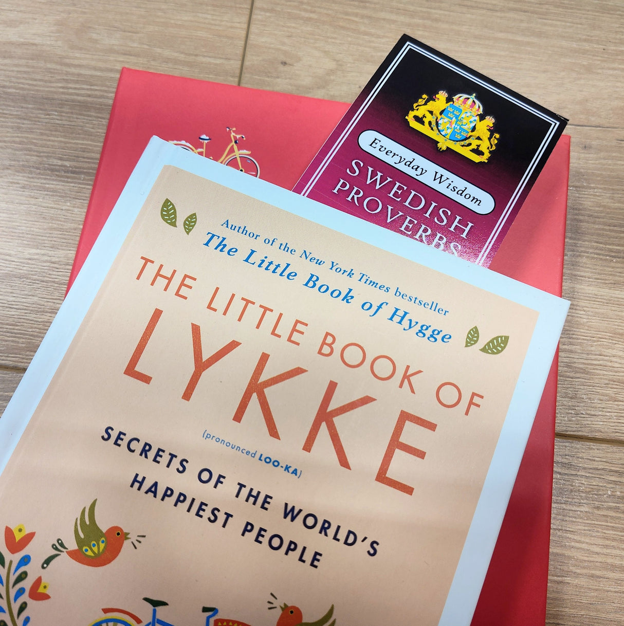 A close-up of The Little Book of Lykke and the Bookmark: Swedish Proverbs “Everyday Wisdom” (2.5" × 8.5") resting on a red surface with a wooden floor background, highlighting Scandinavian tradition.