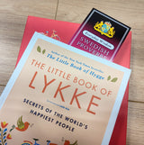 A close-up of The Little Book of Lykke and the Bookmark: Swedish Proverbs “Everyday Wisdom” (2.5" × 8.5") resting on a red surface with a wooden floor background, highlighting Scandinavian tradition.