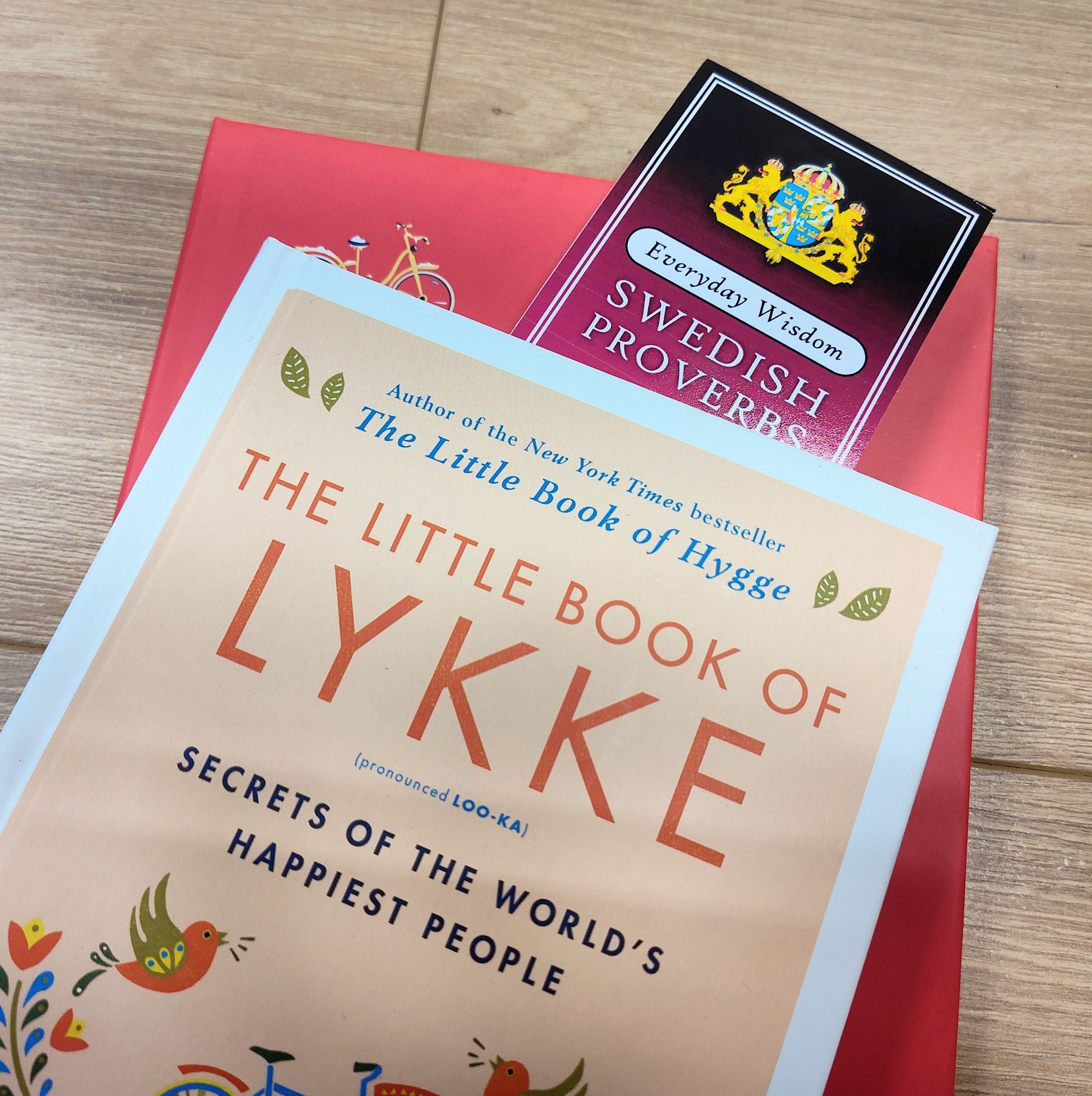 A close-up of The Little Book of Lykke and the Bookmark: Swedish Proverbs “Everyday Wisdom” (2.5" × 8.5") resting on a red surface with a wooden floor background, highlighting Scandinavian tradition.
