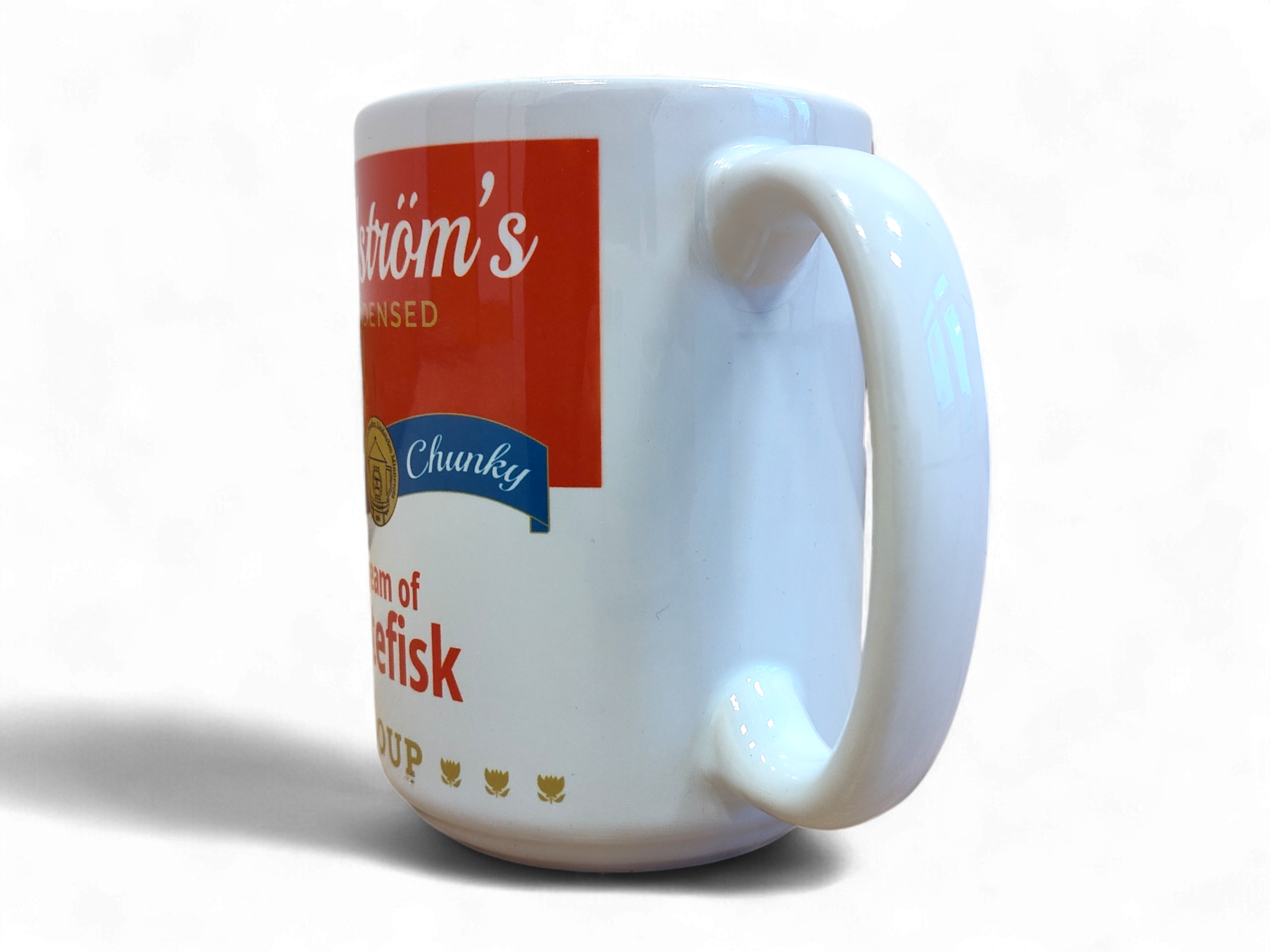 The Lindstrom's Cream of Lutefisk Ceramic Mug (13oz) features a white body with a red and blue label and partial text, shown side-on with the handle visible—a charming Scandinavian gift that celebrates Nordic heritage.