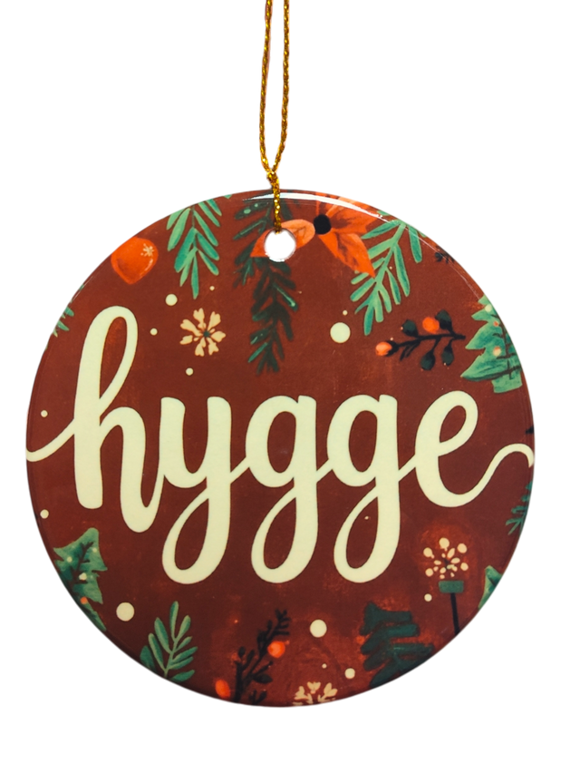 A round ceramic ornament called "Brown Hygge Ceramic Ornament," featuring a brown background and white script, hangs by a gold string—a cozy accent perfect for Scandinavian decor.