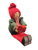 Handcrafted Swedish Santa Boy figurine sits with legs extended, wearing a red hat and green sweater, and holding a small red Dala Horse—perfect for festive Swedish Christmas decor.
