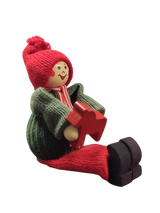 The Handcrafted Swedish Santa Boy Figurine sits holding a red Dala Horse, dressed in a green sweater, red hat, and red pants—perfect for adding Swedish Christmas charm against any neutral white background.