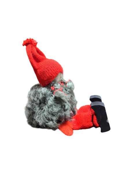The Sitting Santa Girl Figurine features a plush gnome design with a gray beard, red knit hat, mittens, socks, and black shoes. Handmade in Sweden, it’s a charming Christmas décor piece. Shown side view on white background.