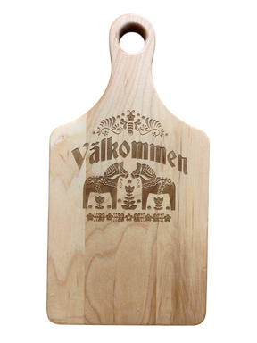 Maple wood cutting board (13.5" x 7") with handle, engraved Swedish folk art design, and “Valkommen” (Welcome in Swedish) at the center.