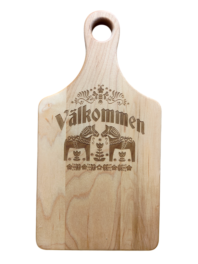 Maple wood cutting board (13.5" x 7") with handle, engraved Swedish folk art design, and “Valkommen” (Welcome in Swedish) at the center.