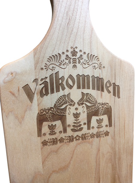 The "Valkommen" Maple Wood Cutting Board (13.5" x 7") features the Swedish word for welcome, two Dala horses, and detailed floral patterns inspired by Swedish folk art.