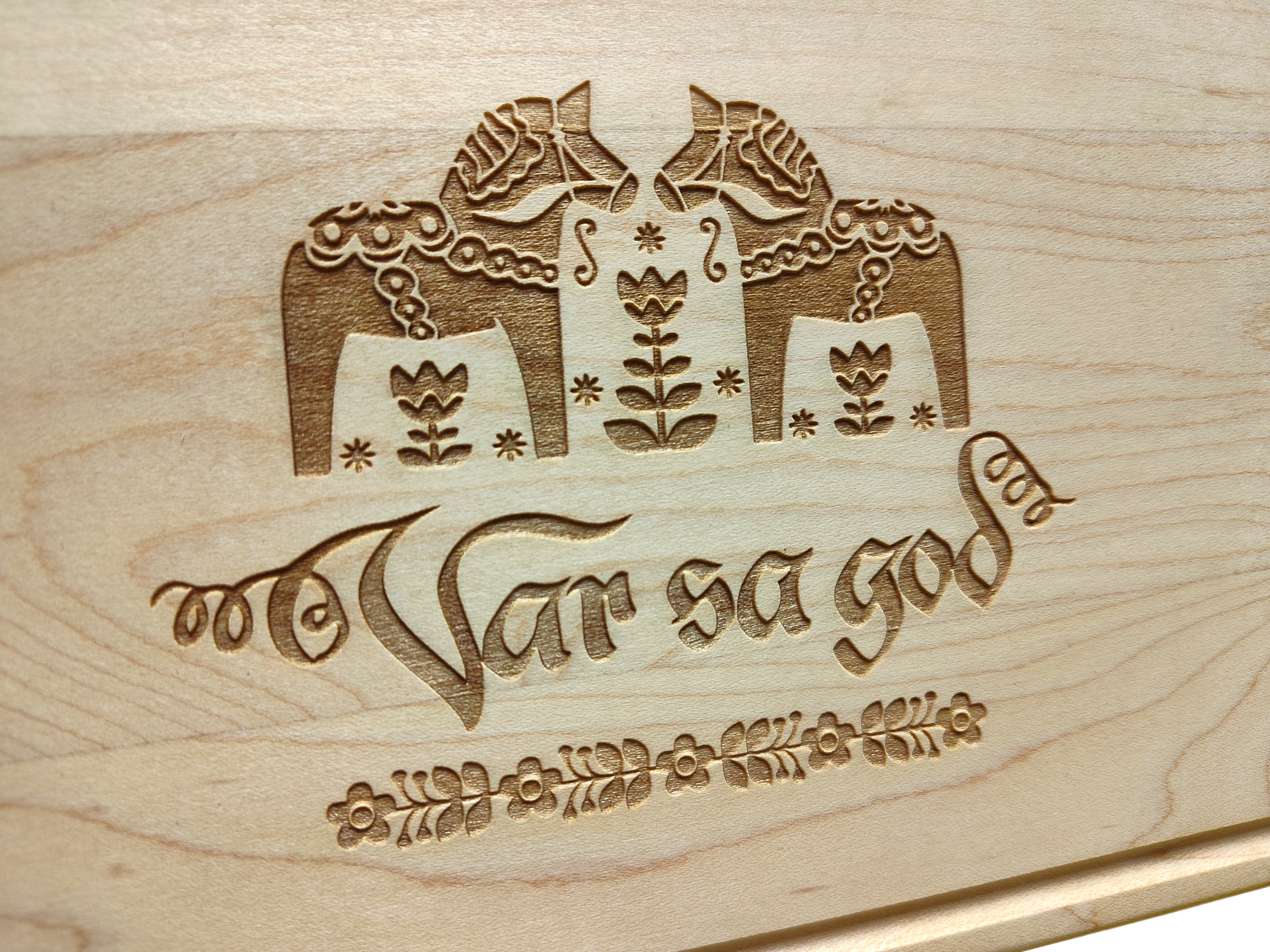 The Var Sa God Maple Cutting Board (9"x6") features two engraved Dala horses, floral designs, and ornate script—ideal as Swedish kitchen decor or unique Scandinavian cutting board art.