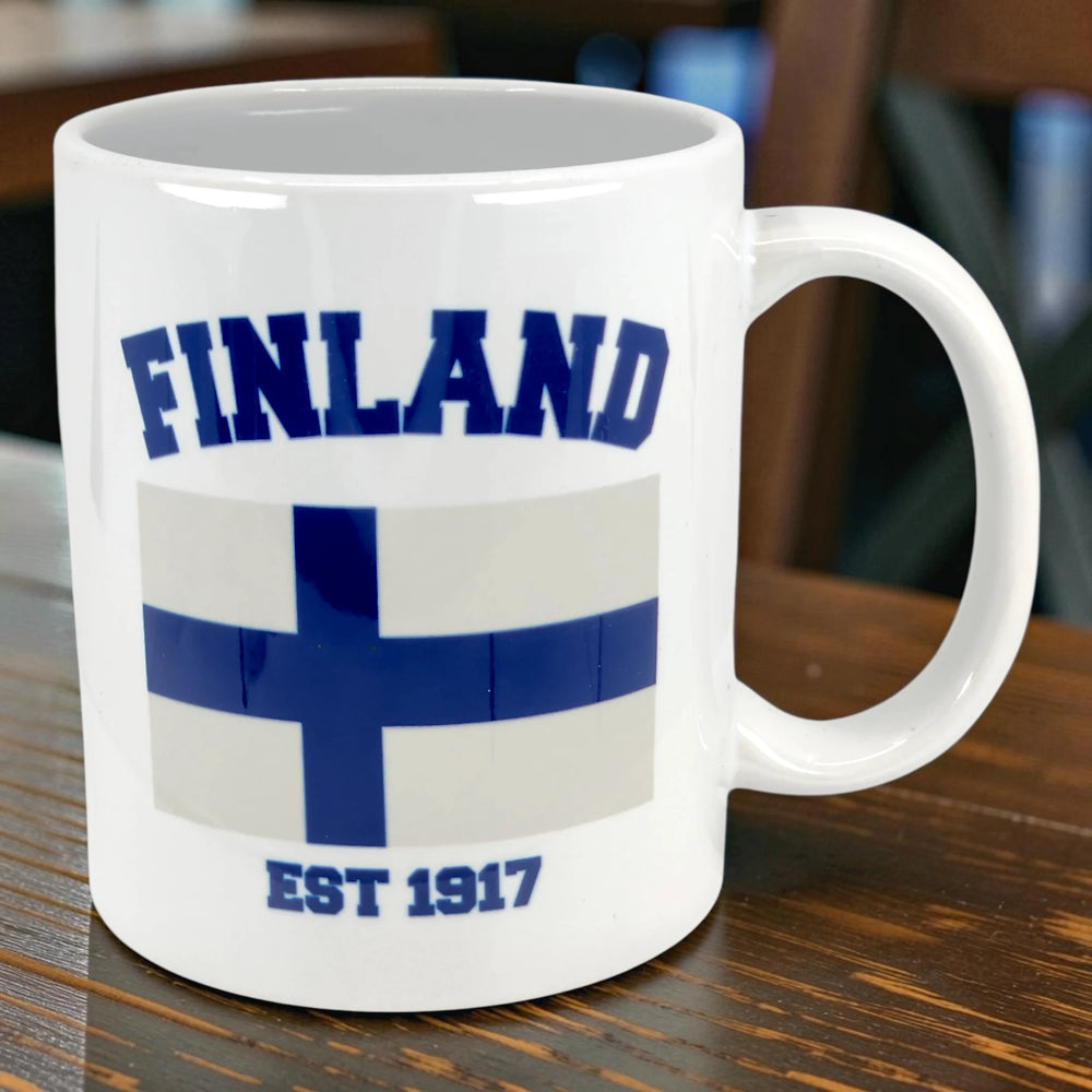 Ceramic coffee mug with the Finland flag design, “Finland” above the blue Nordic cross, and “EST 1917” below, on a wooden surface.