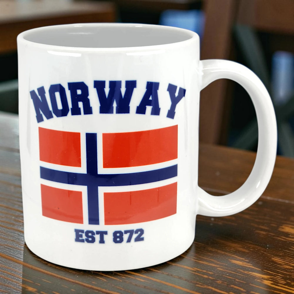 White Scandinavian mug displaying the Norway flag with “NORWAY” above and “EST 872” below, placed on a wooden table.