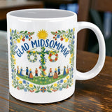 The "Glad Midsommar" Midsummer Mug 11 oz from Swedish Gift Store features colorful flowers, a maypole, and people in traditional dress, with “Glad Midsommar” above the maypole. The mug is shown on a wooden table.