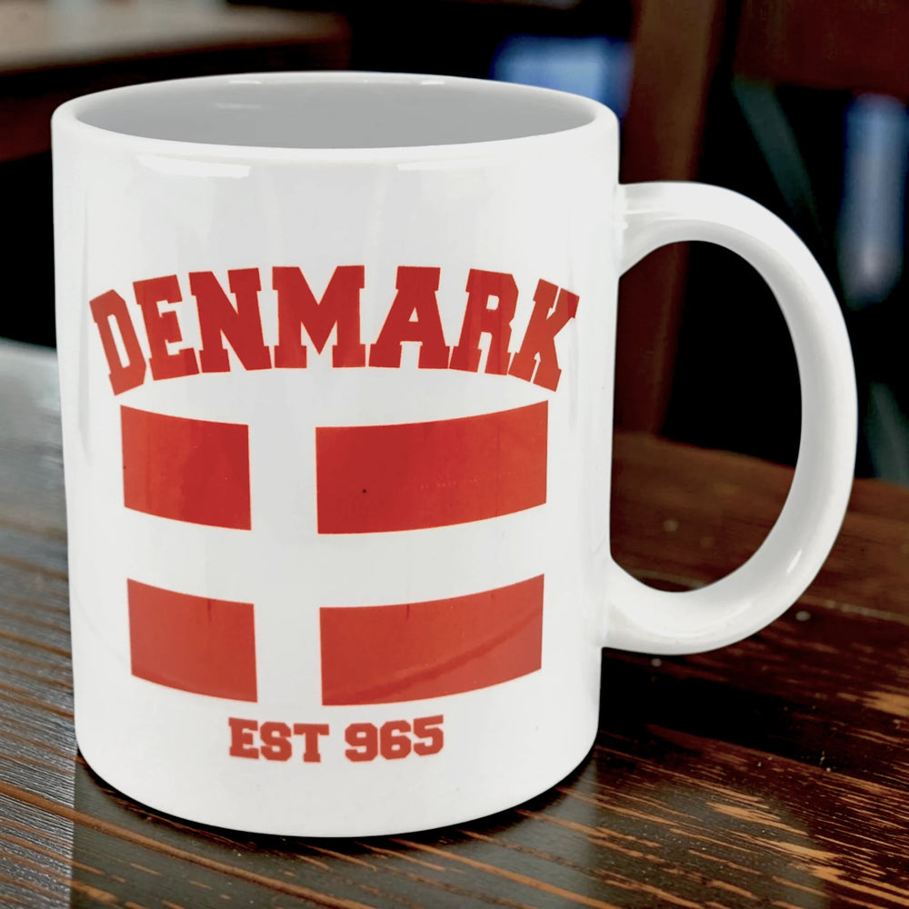 White Danish mug with “DENMARK,” a red flag, and “EST 965” in red lettering, sitting on a wooden surface.