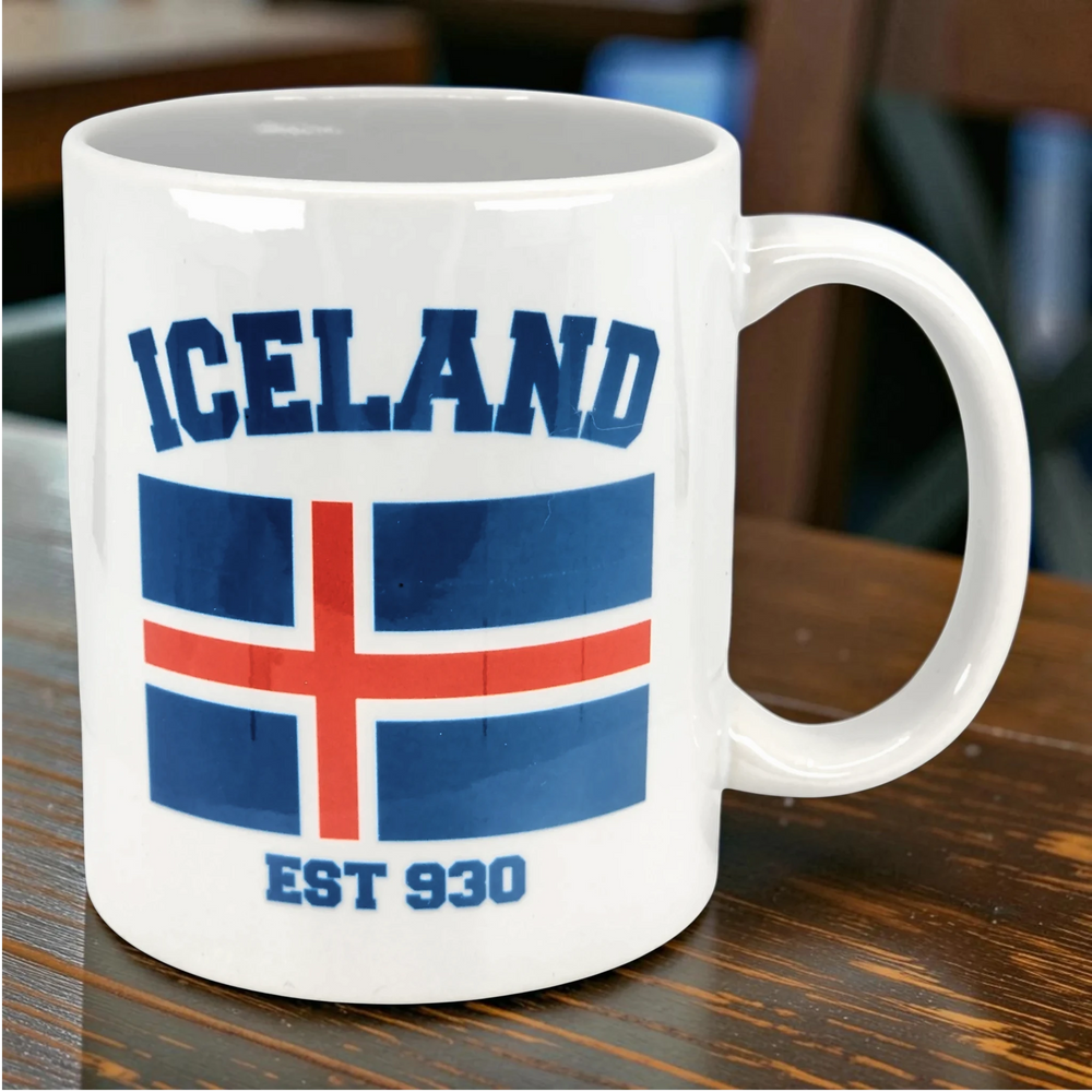 White ceramic mug with “ICELAND,” the Icelandic flag, and “EST 930” in blue and red lettering, placed on a wooden table.