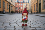 A Scandinavian Girl figurine stands on a cobblestone street in Europe, wearing a red hat and green dress, and holding a handcrafted Dala Horse.