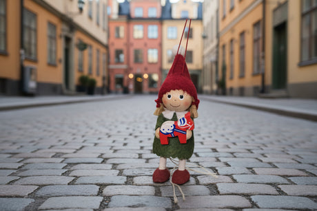 A Scandinavian Girl figurine stands on a cobblestone street in Europe, wearing a red hat and green dress, and holding a handcrafted Dala Horse.