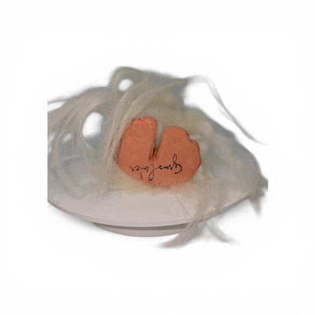 A fragment of terracotta inscribed with writing rests on a white plate, encircled by delicate, pale fibers that evoke the wool and clay materials used in the Gnome: "Foot Tomte"—a gnome with a white hat and clay nose and feet by Gumselids of Sweden, available from Talla Imports—set against a crisp white background.