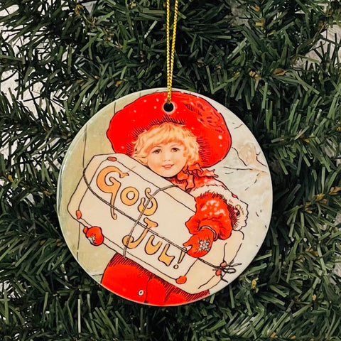 The Ornament: GOD JUL! Girl Holding Sign is a round porcelain piece featuring an illustration of a child in red holding a sign that says "God Jul." It hangs beautifully on a green tree, infusing unique charm into your holiday decoration project.