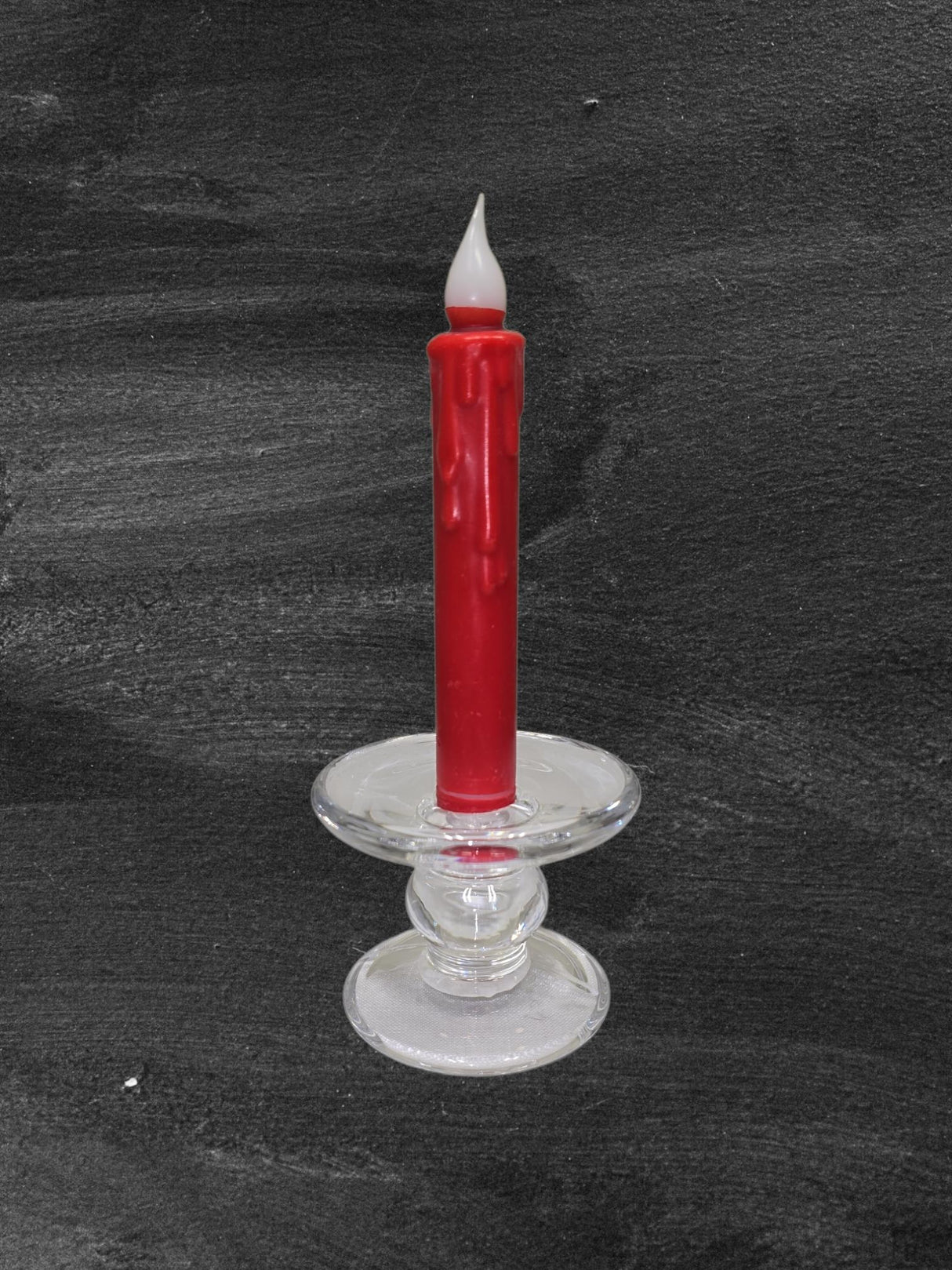 A red wax candle with drips stands in a clear Glass Dual-Size Reversible Candle Holder for taper, ball, or pillar candles, set against a dark textured background.