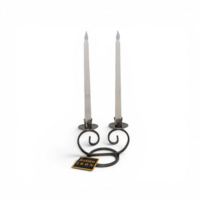 The Talla Imports Danish Iron Double Curls Taper Candle Holder elegantly displays two white taper candles, with a small "DANISH IRON" tag in front, highlighting Scandinavian craftsmanship against a plain white background.