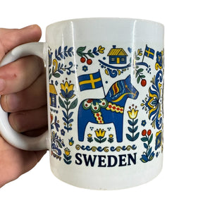 The 11oz ceramic "Mug: Sweden — Swedish Icons & Dala Horse Folk Art" by Swedish Gift Store features blue Dala horses, Swedish flags, flowers, houses, and SWEDEN at the bottom; a hand holds the mug by its handle.