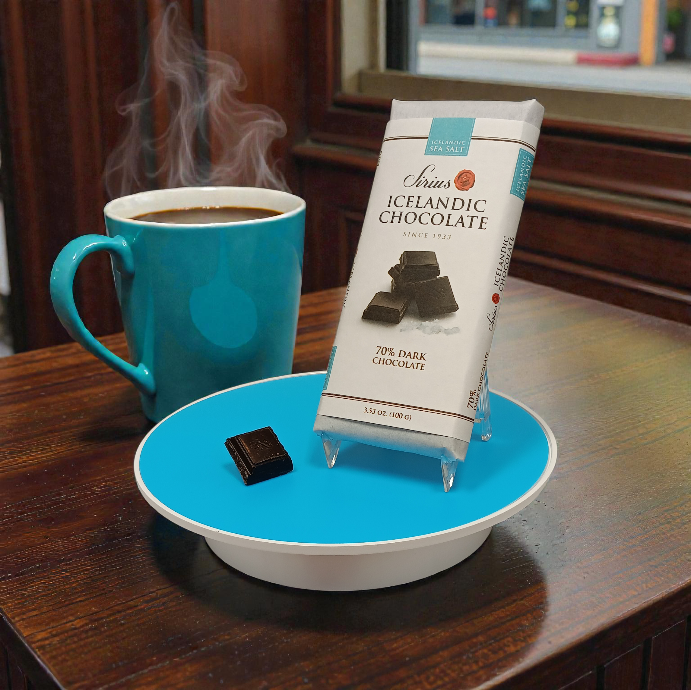 A blue mug of coffee sits on a wooden table next to a plate holding Sirius Icelandic Chocolate—70% Dark Chocolate with Sea Salt (100g)—and a single square of the rich chocolate.