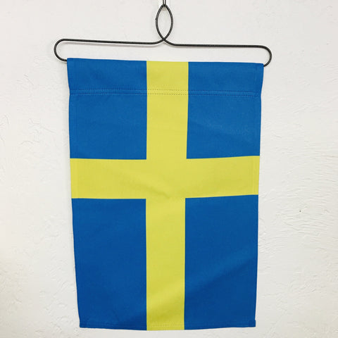 The Garden Flag: Sweden Flag, featuring a blue and yellow design made from durable polyester and equipped with a sewn top pocket for effortless display, hangs elegantly from a wire hanger on a white textured wall.