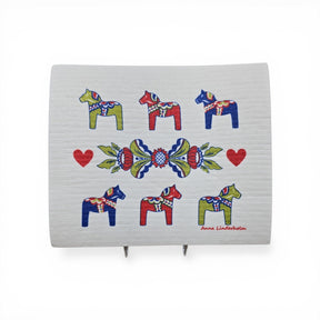 The Dish Cloth: Six Dala Horses with Swedish Kurbits "Flowers" shows six colorful Dala horses, three red hearts, a central floral pattern, and the artist Anna Linderholm's name in the lower right corner.