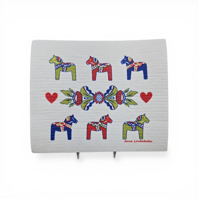 The Dish Cloth: Six Dala Horses with Swedish Kurbits "Flowers" shows six colorful Dala horses, three red hearts, a central floral pattern, and the artist Anna Linderholm's name in the lower right corner.