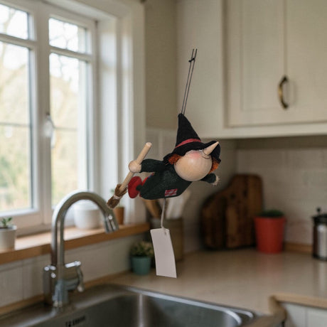 A 4.5" Kitchen Witch Figurine, handcrafted of wood and felt, hangs above the kitchen sink by the window, watching over the plants and kitchen items as a charming household guardian.