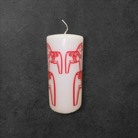 A Logum Kloster Lys Red Dala Horse Pillar Candle from Denmark is shown upright, featuring red line art of dala horses on its white surface against a dark textured background.