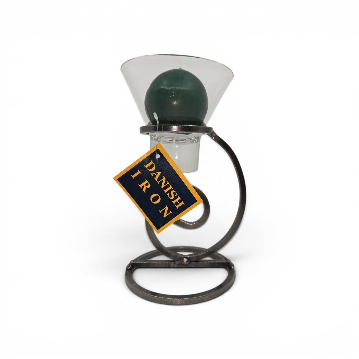 A dark green ball candle in a clear glass Leif cup rests on Talla Imports’ Danish Iron candle holder with a tall curl base; a yellow and blue “Danish Iron” tag hangs from the stand.